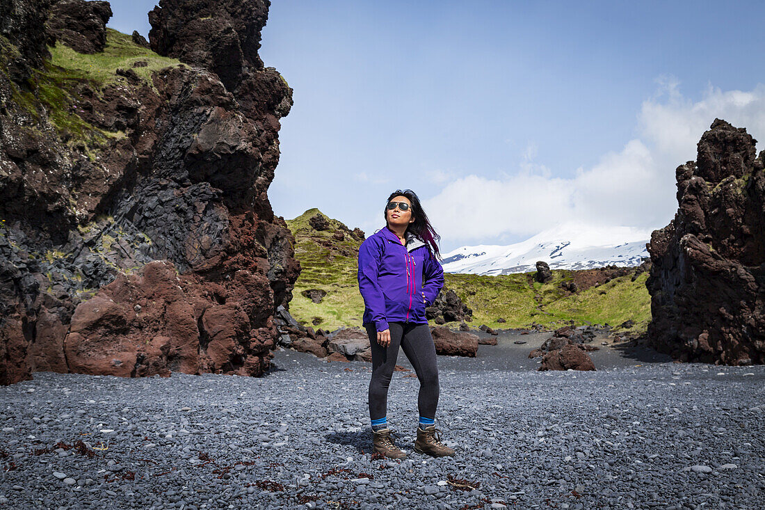 Eine asiatische Touristin posiert für ein Porträt am schwarzen Sandstrand neben einer Felsformation in Westisland, Snaefellsnes-Halbinsel; Island.