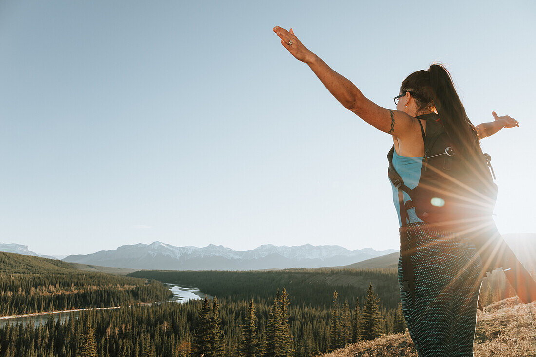 A woman illuminated by sun rays while standing with arms outstretched on a trail in the Rocky Mountains, near Hinton; Alberta, Canada