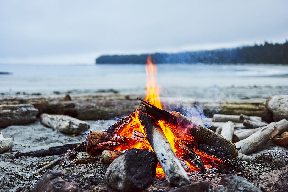 Ein Feuer am Strand mit dem Meer und der Küstenlinie im Hintergrund, Cape Scott Provincial Park; British Columbia, Kanada.