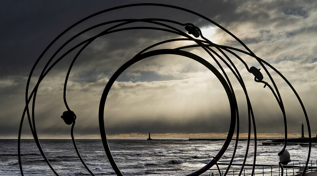 Silhouette einer Skulptur und des Roker-Leuchtturms in der Ferne entlang der Uferlinie des River Ware mit Gewitterwolken über dem Kopf; Sunderland, Tyne and Wear, England.