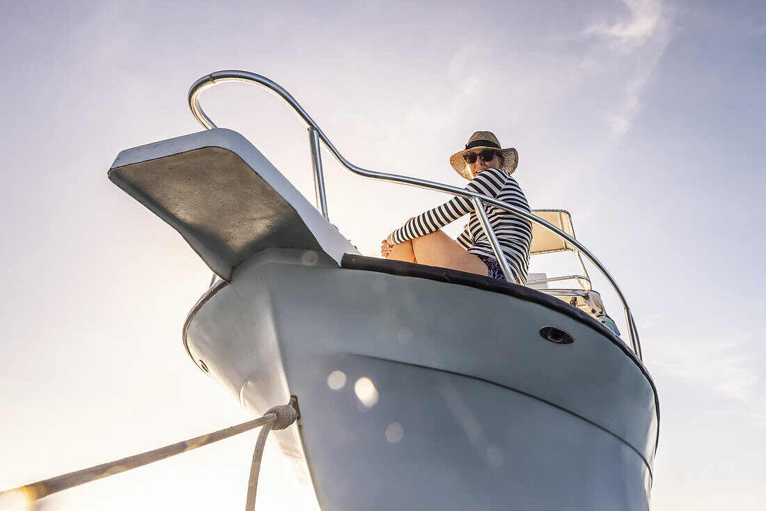 A woman wearing sunglasses and a sun hat looks down at the camera from the deck of a boat against a blue sky with sunlight; Bay Islands Department, Honduras