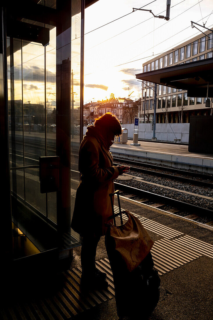 A woman stands with her suitcase on the platform of a train station beside the tracks and uses her smart phone; St. Gallen, St. Gallen, Switzerland