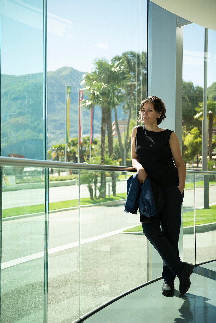 Eine Frau steht an einer Fensterwand und blickt auf die Straße und Palmen; Lugano, Tessin, Schweiz