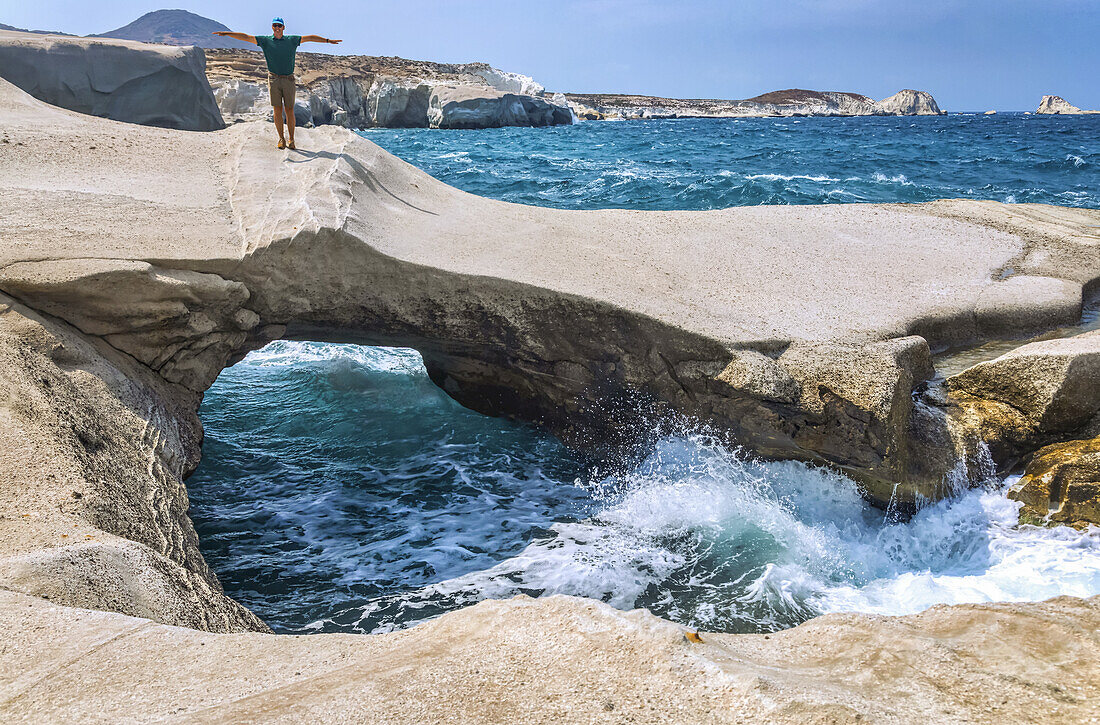 Männlicher Tourist steht auf einzigartiger Felsformation mit natürlichem Bogen und Brücke entlang der Küste von Milos, einer griechischen Insel; Milos, Griechenland