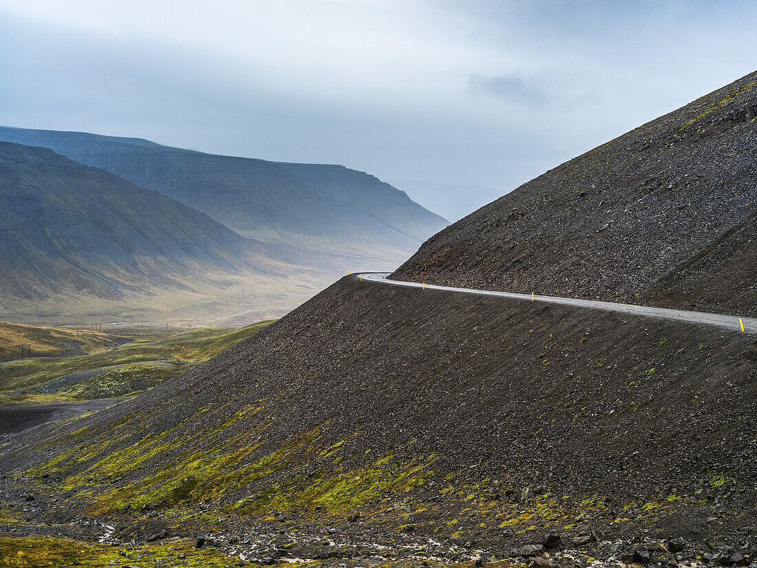 Straße, die um eine Kurve an einem Hang führt, mit Blick auf ein Tal und Berge unter einem bedeckten Himmel; Westfjorde, Island