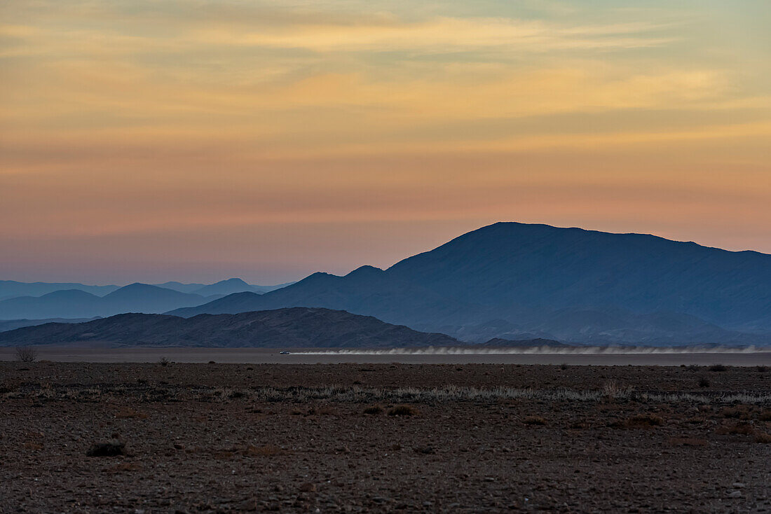 Sonnenaufgang in Aluvlei, Namib-Naukluft-Nationalpark, mit einem Auto, das über die Landschaft fährt und Staub in der Luft hinterlässt; Namibia.
