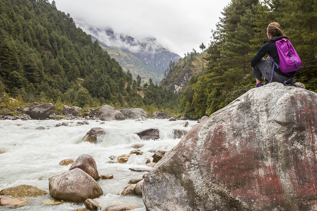 Kaukasische Frau mit rosa Rucksack sitzt an einem späten Herbstnachmittag auf einem großen Felsbrocken am Rande des Dudh Koshi-Flusses in Phakding, Solokhumbu-Distrikt, Nepal; Phakding, Solokhumbu-Distrikt, Nepal