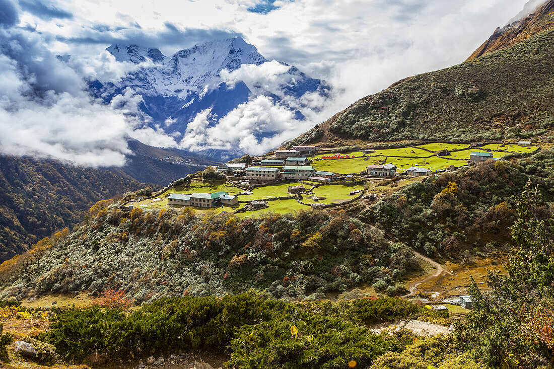 Village of Dole, in the Solokhumbu district, Sagarmatha National Park, Nepal, is lit up by the sun as it sits on a green terrace high up in the Himalayan mountains along the Gokyo trek trail, while mountains covered with fresh snow peak out from the clouds in the distance; Dole, Khumbu Region, Nepal