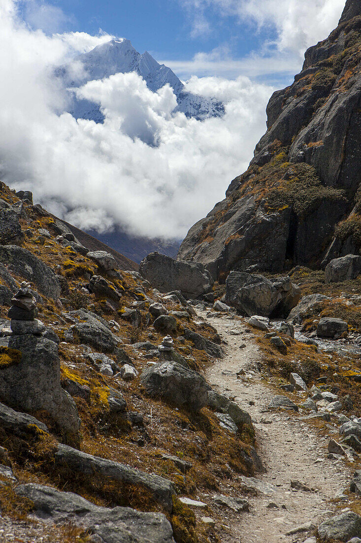 Gokyo-Trek mit herbstlich gefärbter Tundra und Steinhaufen am Wegesrand, während Wolken den schneebedeckten Himalaya-Gipfel im Hintergrund an einem sonnigen Tag in der Nähe von Gokyo, Sagarmatha-Nationalpark, Solokhumbu-Distrikt, Nepal, teilweise verdecken.