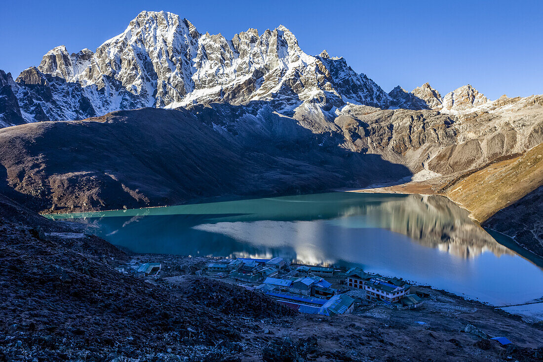 Village of Gokyo in shadow is dwarfed by the surrounding Himalayan Peaks with the reflection of the snow covered glacial moraine of the Ngozumpa Glacier in the turquoise colored Gokyo Lake on a crisp autumn morning; Sagarmatha National Park, Solokhumbu District, Nepal