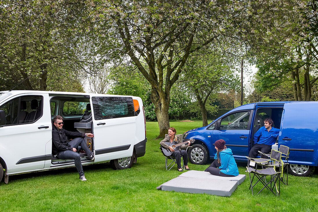 Friends relaxing while setting up camp outside two modified caravan campers; Bourton-on-the-Water, England, United Kingdom