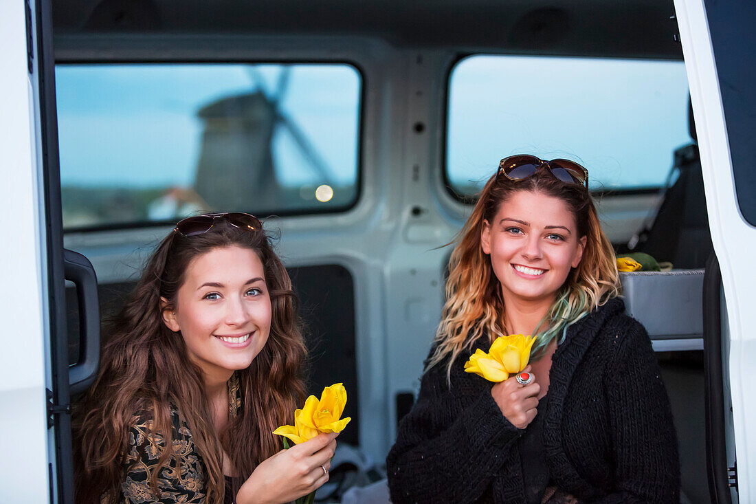 Zwei junge Frauen sitzen in einem Fahrzeug und machen Urlaub mit frisch gepflückten gelben Tulpen. Eine Fahrt außerhalb von Amsterdam Central führt in die Küstenstadt Egmond aan Zee. Reisende machen sich auf die Suche nach Hollands berühmten Windmühlen; Egmond aan Zee, Holland