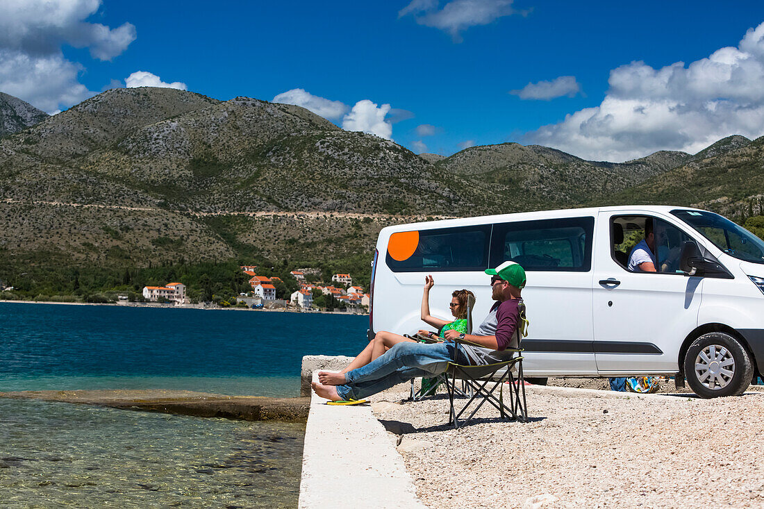 Freunde sitzen in der Sonne und ruhen sich vor einem Wohnmobil aus, als eine Gruppe von Reisenden in Slano für einen Nachmittag am Strand anhält; Slano, Gespanschaft Dubrovnik-Neretva (Dubrova?ko-neretvanska ?upanija), Kroatien
