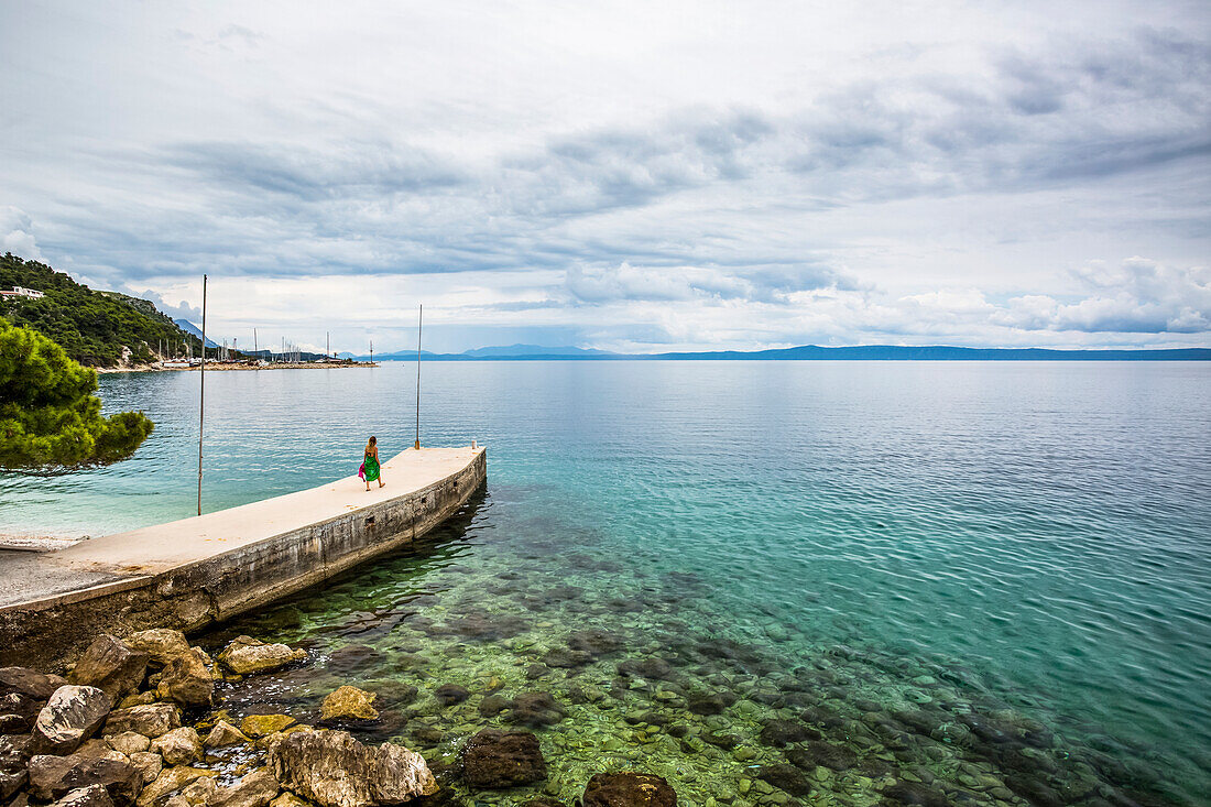 A young woman walks on a pier towards the turquoise water along the coast of Podgora, Croatia. A group stops for an early morning swim and shower after camping in their van; Podgora, Split-Dalmatia County, Croatia