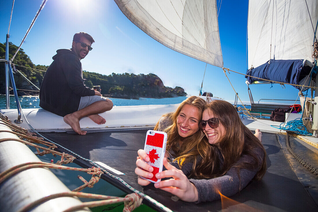 Friends taking a selfie onboard a catamaran boat tour through Abel Tasman National Park with a man smiling in the background; Tasman, New Zealand