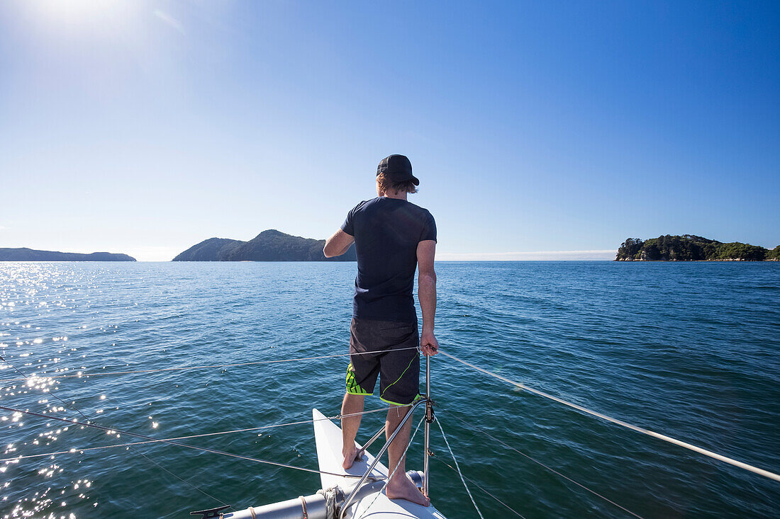 Blick von hinten auf einen Mann, der auf einer Bootstour im Abel-Tasman-Nationalpark am Bug eines Katamarans steht; Tasman, Neuseeland.