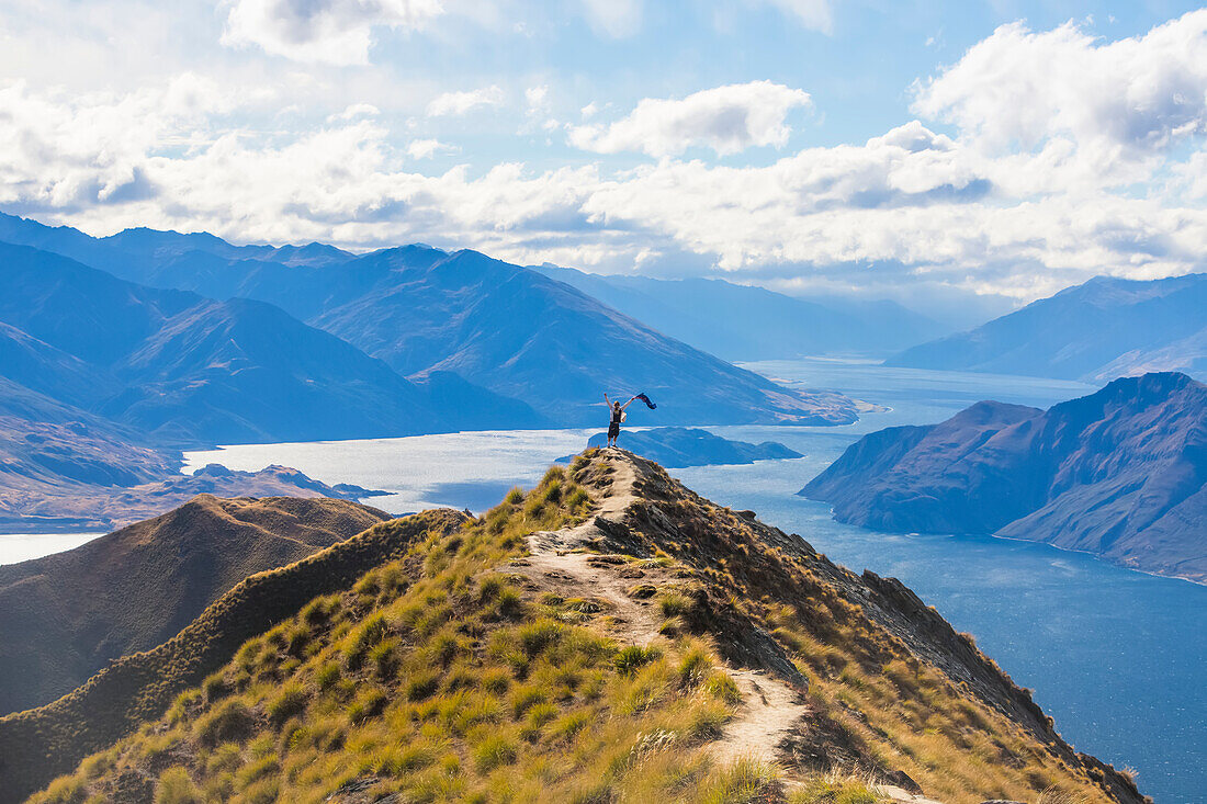 Mann steht auf dem Gipfel des Roys Peak und schwenkt die neuseeländische Flagge nach der anstrengenden Wanderung zum Aussichtspunkt, von dem aus man einen spektakulären Blick auf den Wanaka Lake und die umliegenden Berge der Südalpen hat; Otago, Neuseeland