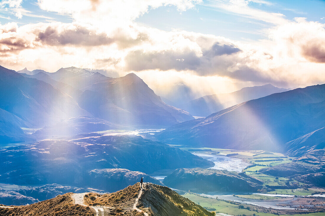 Mann steht auf dem Gipfel des Roys Peak und genießt die herrlichen Sonnenstrahlen nach der anstrengenden Wanderung zum Aussichtspunkt, von dem aus man einen spektakulären Blick auf die Seen und die umliegenden Berge der Südalpen bei Wanaka hat; Otago, Neuseeland