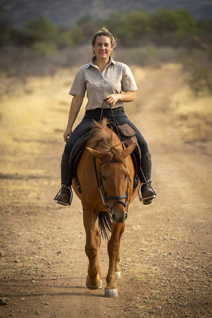 Woman riding horse (Equus ferus caballus) on a dirt road through the bush on the savanna looking at the camera and smiling at the Gabus Game Ranch; Otavi, Otjozondjupa, Namibia