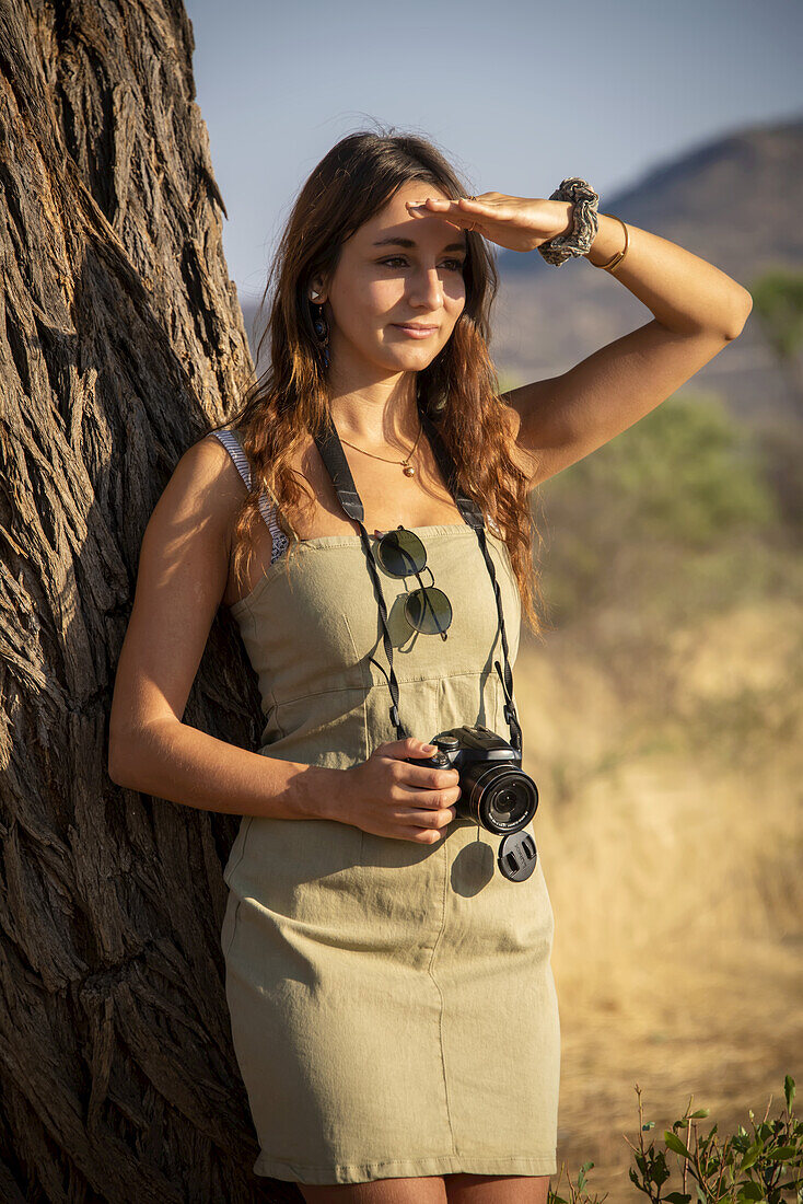 Porträt einer Frau, die sich an einen Baumstamm lehnt, ein Sonnenkleid trägt und eine Kamera in der Hand hält, während sie ihre Augen gegen das helle Sonnenlicht abschirmt und in die Ferne blickt, auf der Gabus Game Ranch; Otavi, Otjozondjupa, Namibia.
