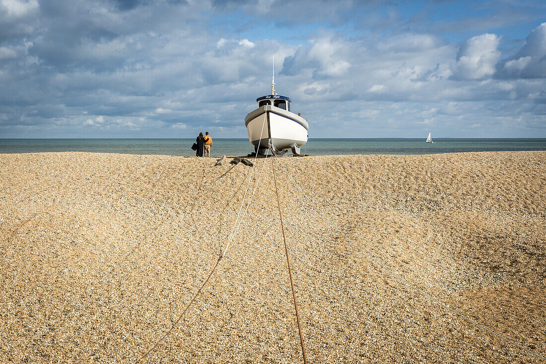 Couple stands at the water's edge looking out into the ocean next to a wooden cabin boat tethered in the middle of the shingle beach at Dungeness along the Atlantic Coast; Dungeness, Kent, England, United Kingdom