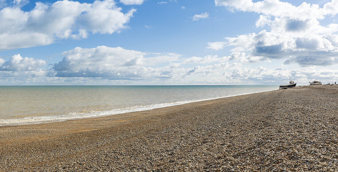 View of the shoreline along the Atlantic Ocean at Dungeness shingle beach on a sunny day with a beached cabin boat in the distance; Dungeness, Kent, England, United Kingdom