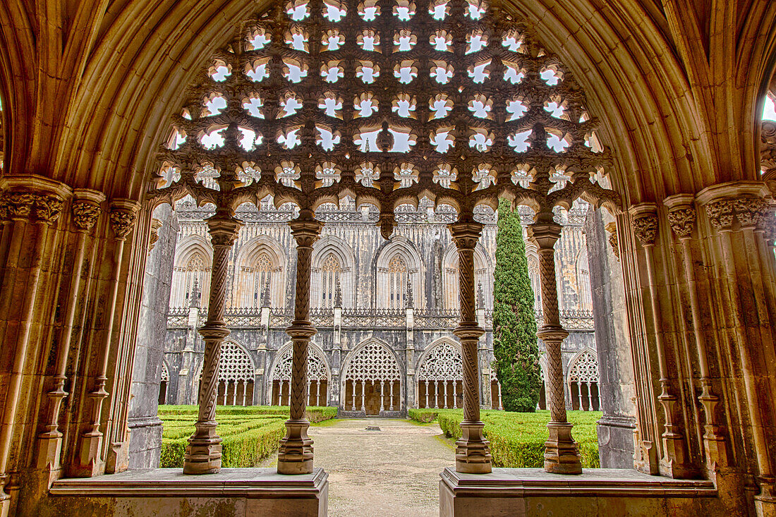 Looking through the ornate columns and archway of the cloister into the formal garden and walkway of the inner courtyard towards the chapel at the medieval Monastery of Batalha, a masterpiece of Gothic architecture; Batalha, District of Leiria, Centro Region, Portugal