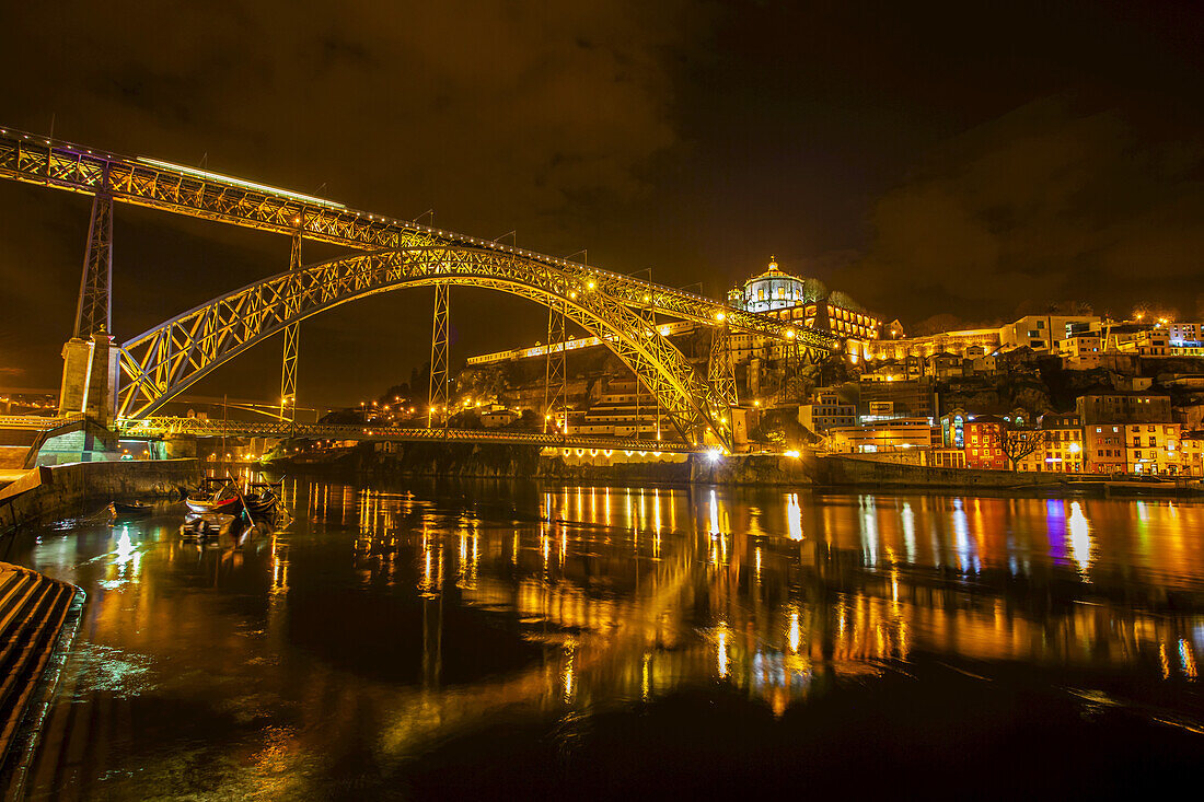 Dom-Luis-I-Brücke, die den Fluss Douro zwischen den Städten Porto und Vila Nova de Gaia überspannt, mit Blick auf das Kloster aus dem 17. Jahrhundert, das Mosteiro da Serra de Pilar und die Hafenlagerhäuser von Vila Nova de Gaia, die bei Nacht beleuchtet sind; Porto, Region Norte, Portugal.