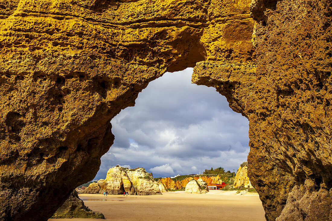 Looking through the detail of a sea arch on Dona Ana Beach at low tide with its sunlit shoreline and sandstone sea stacks along the famous Algarve coastline under a stormy sky; Praia da Dona Ana, Lagos, Algarve Region, Portugal