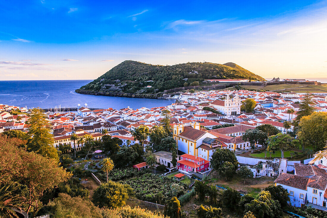 Blick auf den Monte Brasil und die Hauptstadt Angra do Heroismo, mit dem Sonnenuntergang, der warmes Licht auf die weiß getünchten Gebäude und den strahlend blauen Atlantik wirft; Terceira, Azoren
