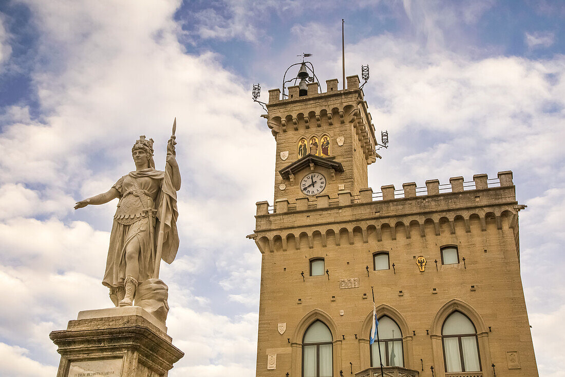 Piazza della Liberta in der Stadt San Marino mit dem Dach des neugotischen Rathauses und einer Marmorskulptur einer weiblichen Figur, die die Freiheit darstellt (Statua della Liberta), vor einem wolkenverhangenen blauen Himmel; Republik San Marino, Nordmittelitalien.