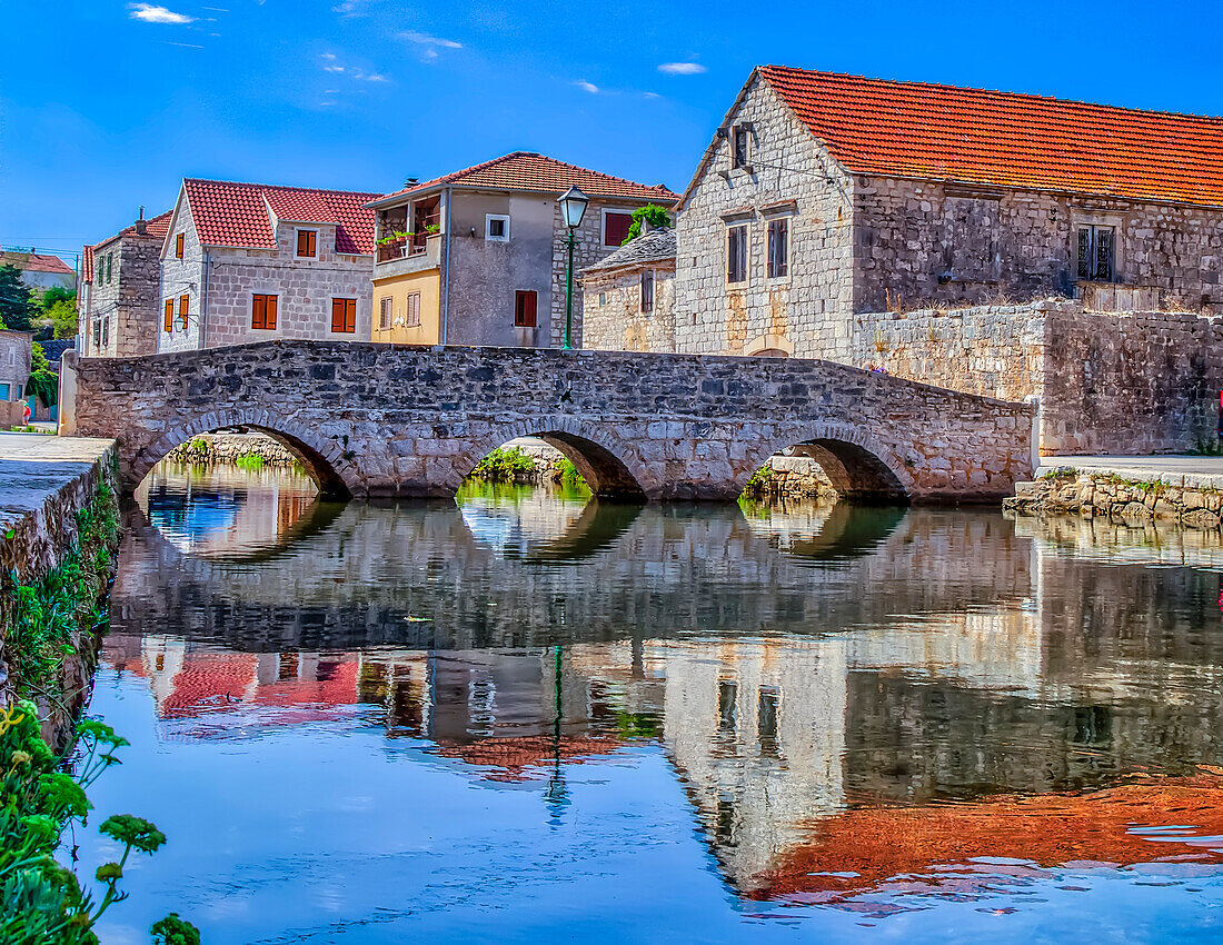 Mittelalterliche Bogenbrücke über einen Kanal mit alten Steinbauten, die sich im Wasser spiegeln; Vrboska, Insel Hvar, Kroatien.