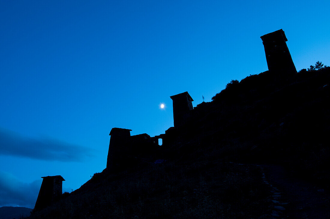 Dämmerung über den mittelalterlichen Festungstürmen von Keselo, die die Silhouette des Berghangs vor dem Hintergrund des Mondes im Tuscheti-Nationalpark zeichnen; Omalo, Kachetien, Georgien.