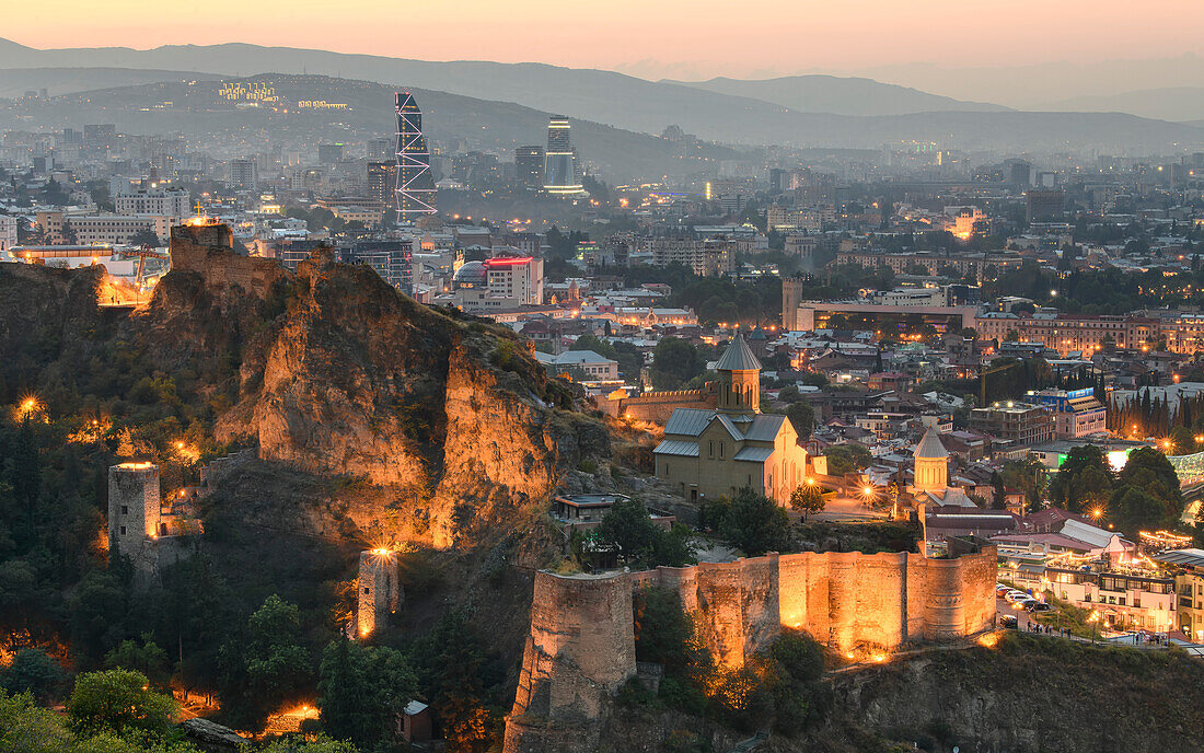 Overview of the capital city with the illuminated, ancient walled fortress of Narikala situated on the hillside above the Kura (Mtkvari River) with its 20th century restoration of St Nicholas Church on the lower court, at dusk; Tbilisi, Georgia