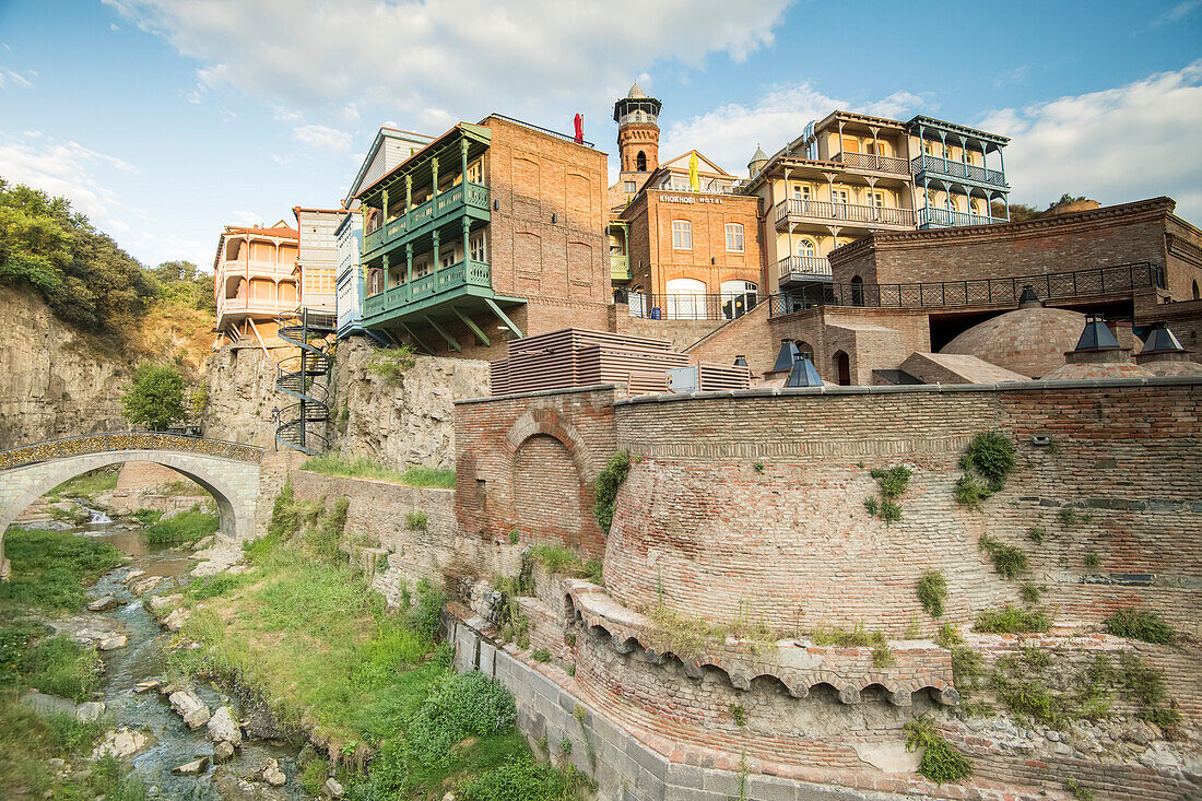 A small arch bridge crossing the Tsavkisi-Tskali River with love locks attached to its railings in front of the old buildings on the cliffs in Legvtakhevi, part of the historical neighborhood of Abanotubani in the Old Town with the domes of the hot sulfur bath houses above the retaining wall and the minaret of the Tbilisi Mosque in the background; Tbilisi, Georgia