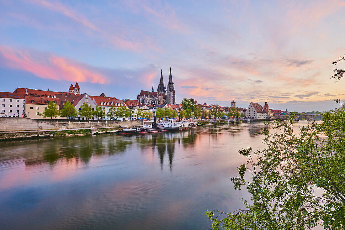 Outlook over the Danube River with the old, 12th Century Stone Bridge in the distance and the Gothic St Peter's Cathedral from the Marc?-Aurel-shore in the Old Town of Regensburg at sunset with a pastel hues; Regensburg, Bavaria, Germany