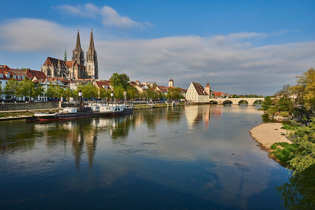 Outlook over the Danube River with the old, 12th Century Stone Bridge in the distance and the Gothic St Peter's Cathedral from the Marc?-Aurel-shore in the Old Town of Regensburg with a blue sky at sunset; Regensburg, Bavaria, Germany