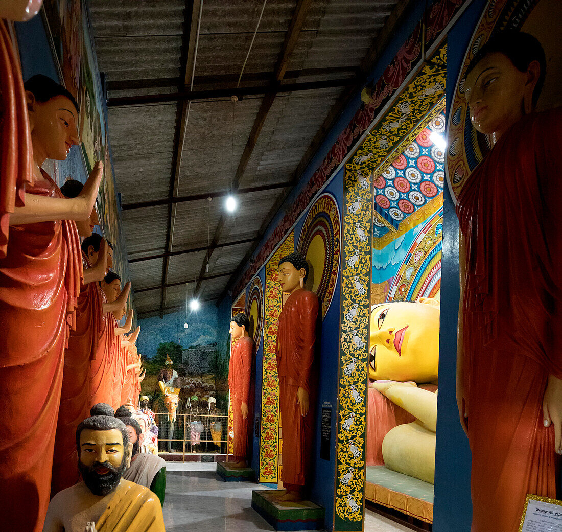 Flur gesäumt von Statuen stehender Buddhas und Skulpturen heiliger Männer, die vor ihnen sitzen, mit dem längsten liegenden Buddha Südasiens, der durch die Öffnungen in der Wand im buddhistischen Kloster Galagoda Shailatharama Viharaya zu sehen ist; Balapitiya, Distrikt Galle, Sri Lanka.