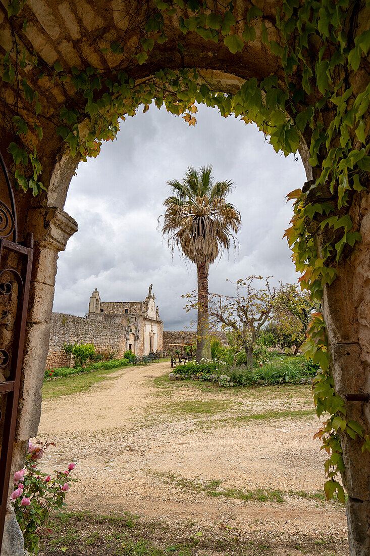 Blick durch einen Torbogen auf einen abgelegenen Garten mit grauen Wolken über einer italienischen Villa in den Hügeln über Ostuni; Ostuni, Apulien, Italien.