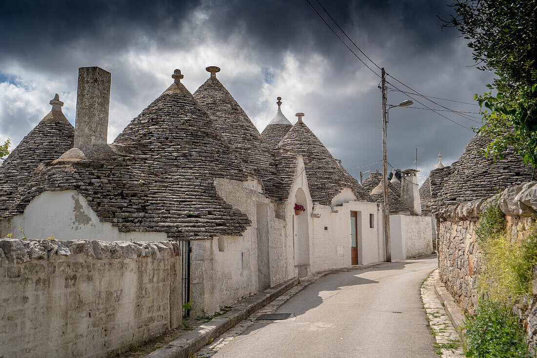 Straßenszene mit einer Reihe traditioneller apulischer Trulli-Häuser aus rundem Stein vor einem stürmischen Himmel an einem sonnigen Tag in der Stadt Alberobello; Alberobello, Apulien, Italien.