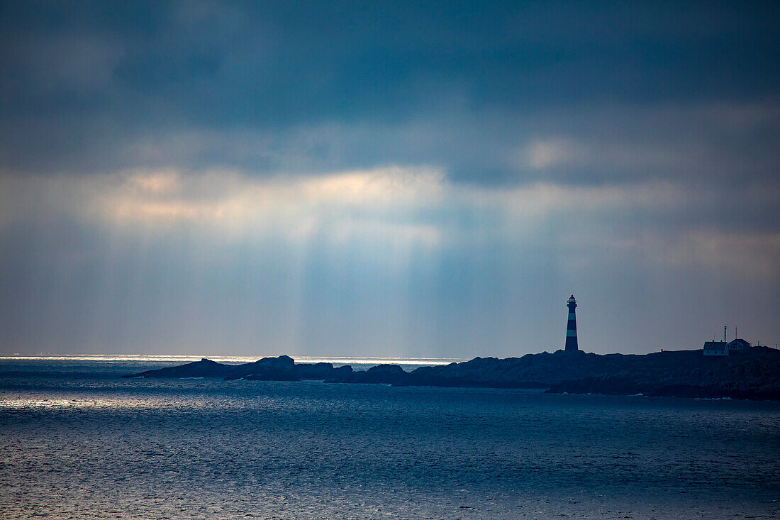 Hellisoy-Leuchtturm, ein abgelegener Leuchtturm auf einer Insel in den westlichen Fjorden Norwegens mit Sonnenstrahlen, die sich in einem stürmischen Himmel spiegeln; Hordaland, Norwegen
