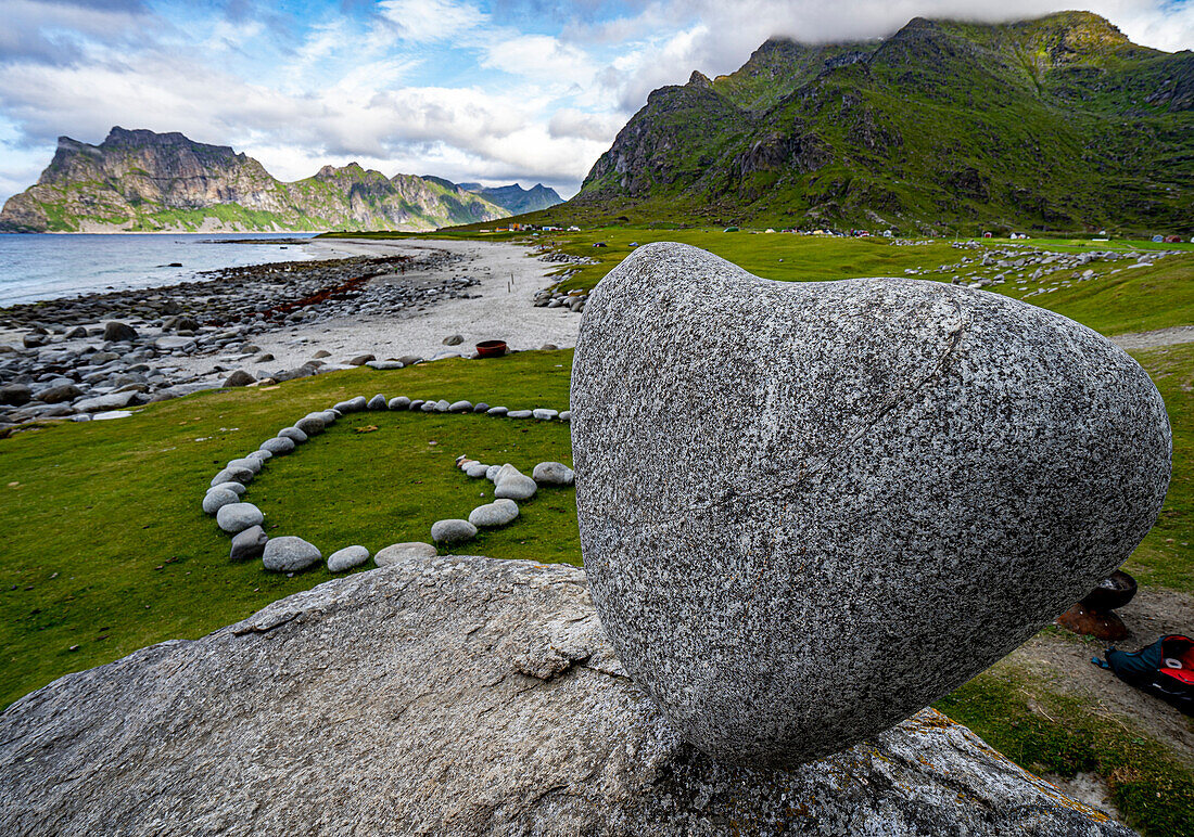 Die dramatische Küstenberg- und Felslandschaft des Uttakleiv-Strandes auf den Lofoten, Norwegen, mit dem natürlich geformten herzförmigen Felsen, der auf einem großen Felsblock balanciert, und anderen Felsen, die in einem herzförmigen Muster auf dem Gras liegen; Lofoten, Norwegen.