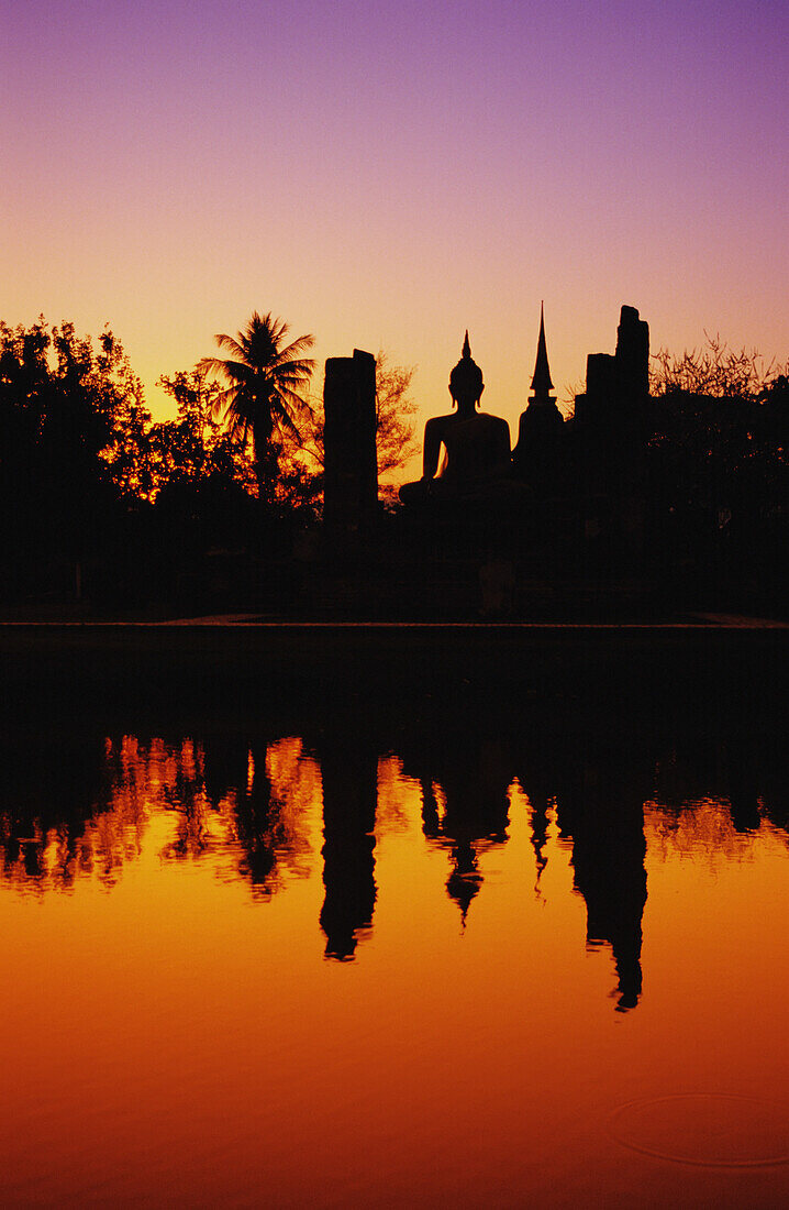 Thailand, Sukhothai, Wat Mahathat, Distant View Of Buddha Statue At Sunset, Silhouetted And Reflecting On Water.