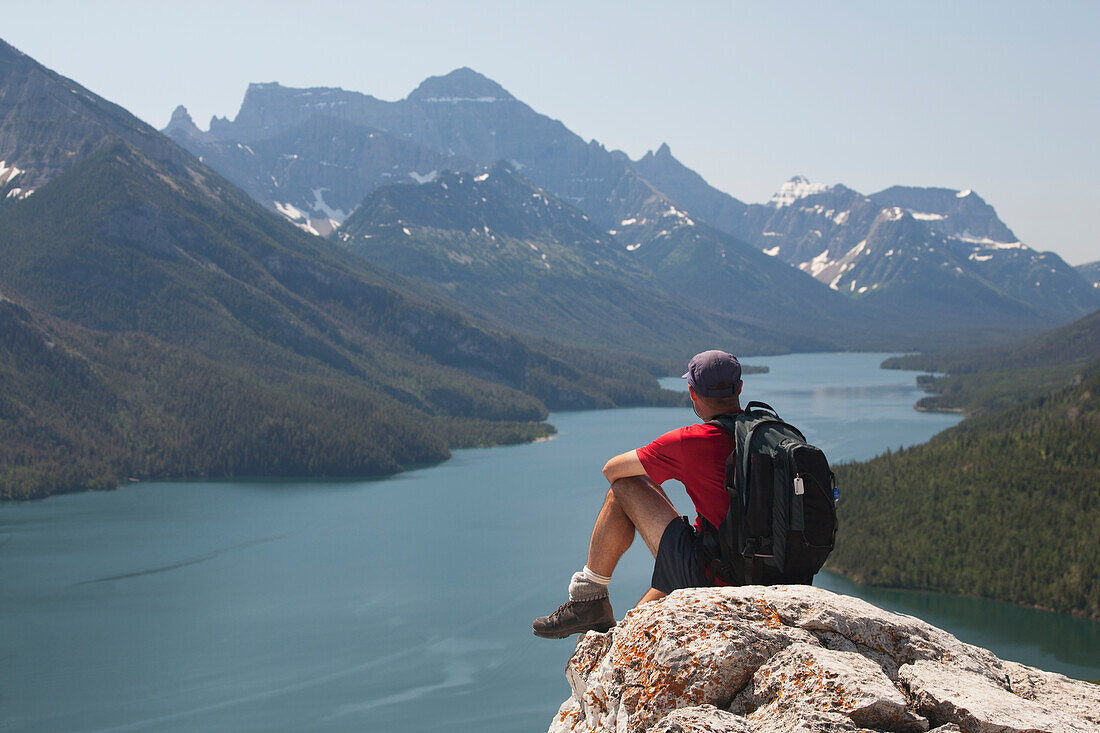 Männlicher Wanderer mit Rucksack auf einer Felsklippe sitzend mit Blick auf einen See mit Bergen und blauem Himmel; Waterton Alberta Kanada