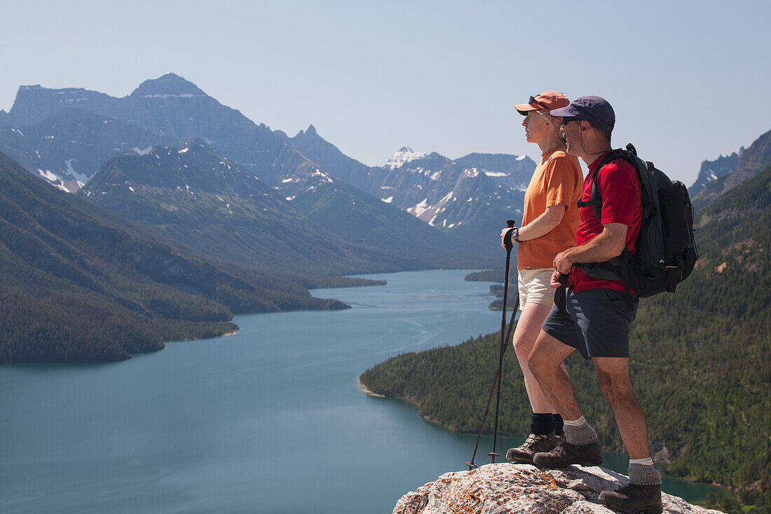 Male And Female Hiker Standing On A Rock Cliff Looking Out Onto A Lake With Mountains And Blue Sky; Waterton Alberta Canada