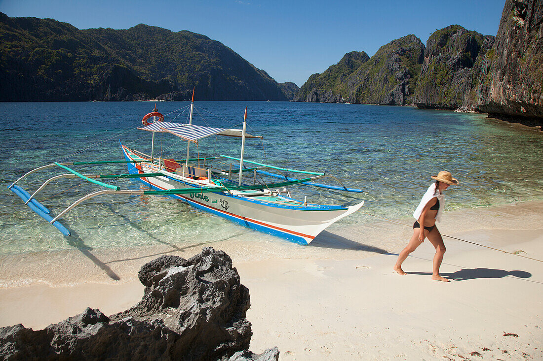 Eine Touristin mit Sonnenhut und Bikini geht an einem Bangka-Boot auf der Insel Matinloc bei El Nido und Corong Corong vorbei; Bacuit-Archipel Palawan Philippinen