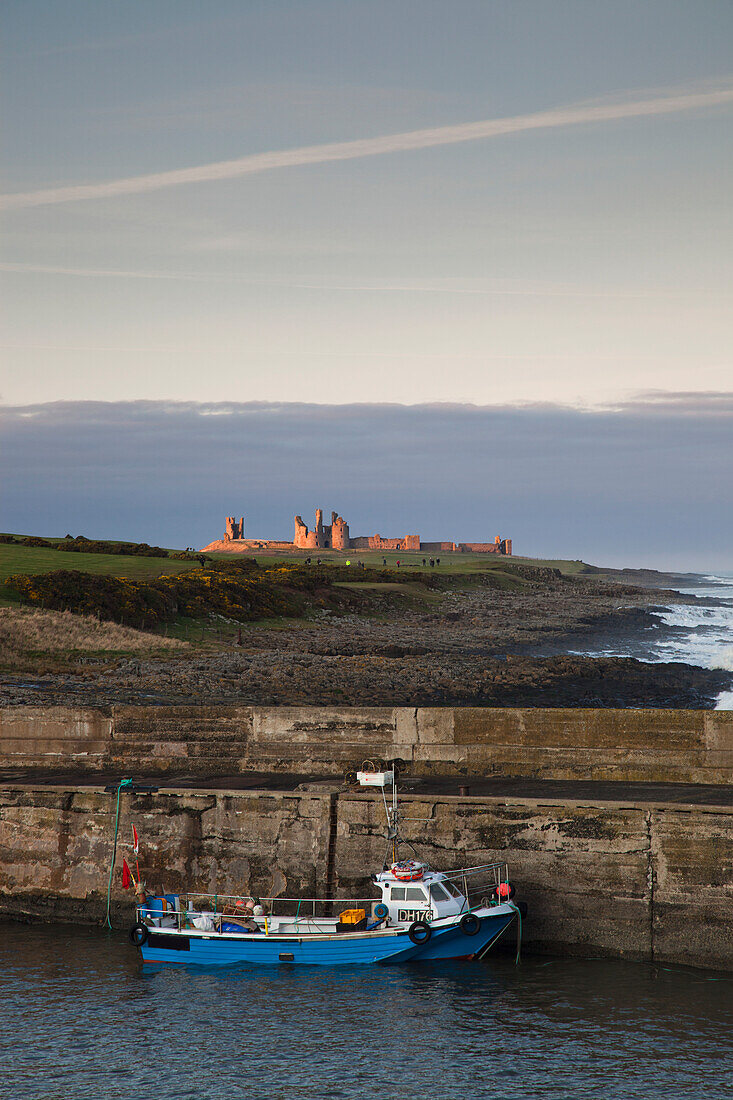 A Fishing Boat Moored Along A Stone Wall On The Coast With Dunstanburgh Castle In The Distance; Northumberland England