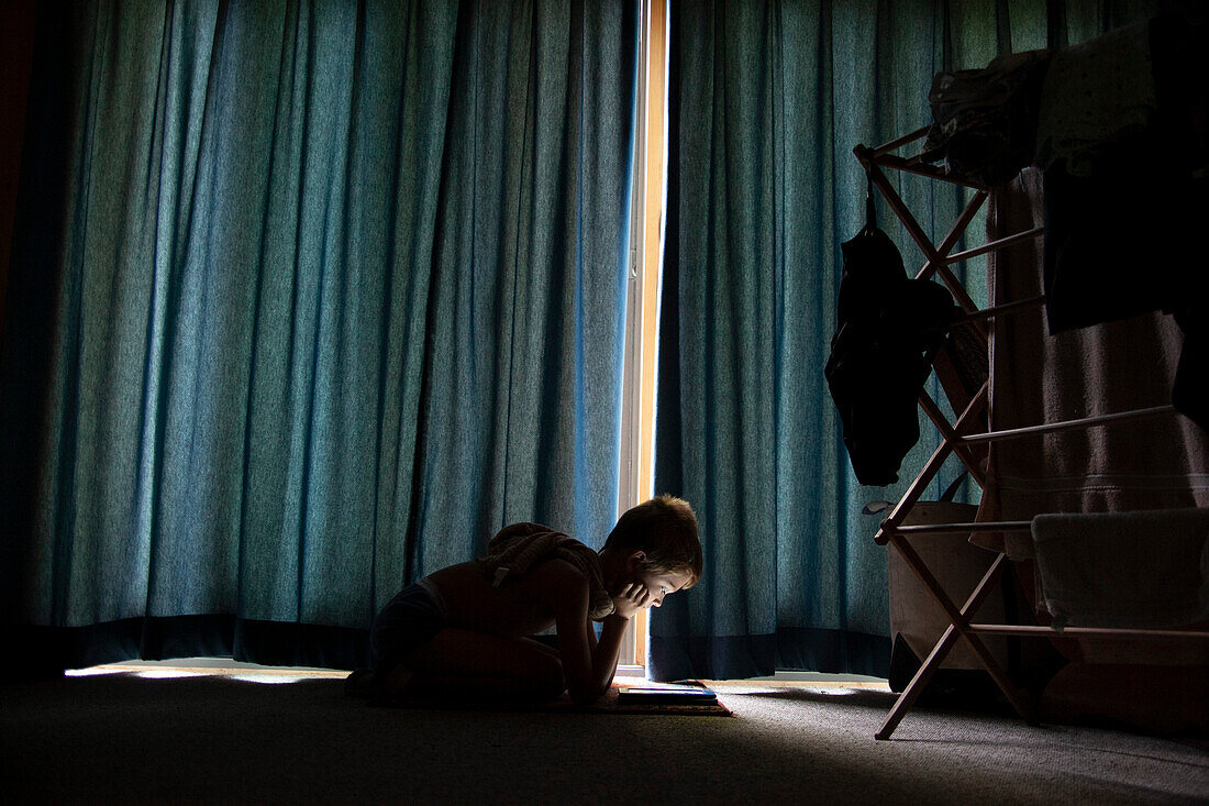 Junge sitzt auf dem Boden und liest im Sonnenlicht, das durch die geschlossenen Vorhänge dringt