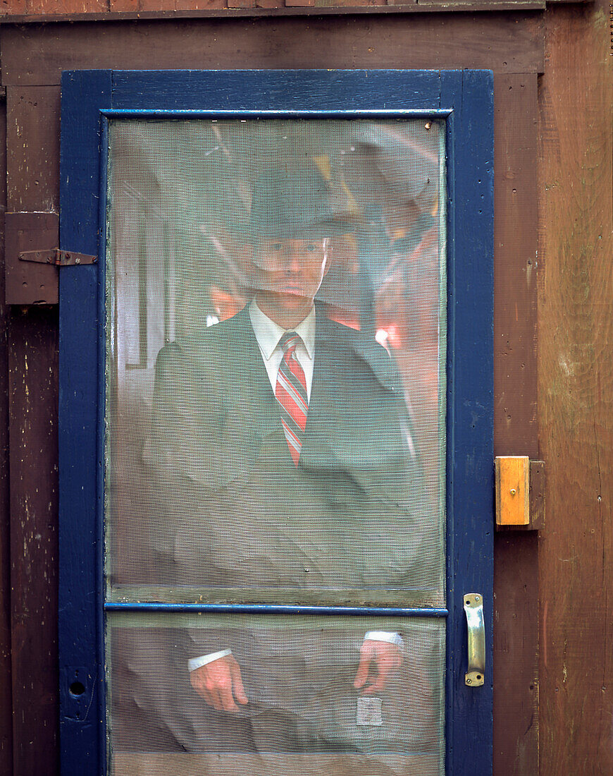 Man in suit and hat standing behind rustic screen door
