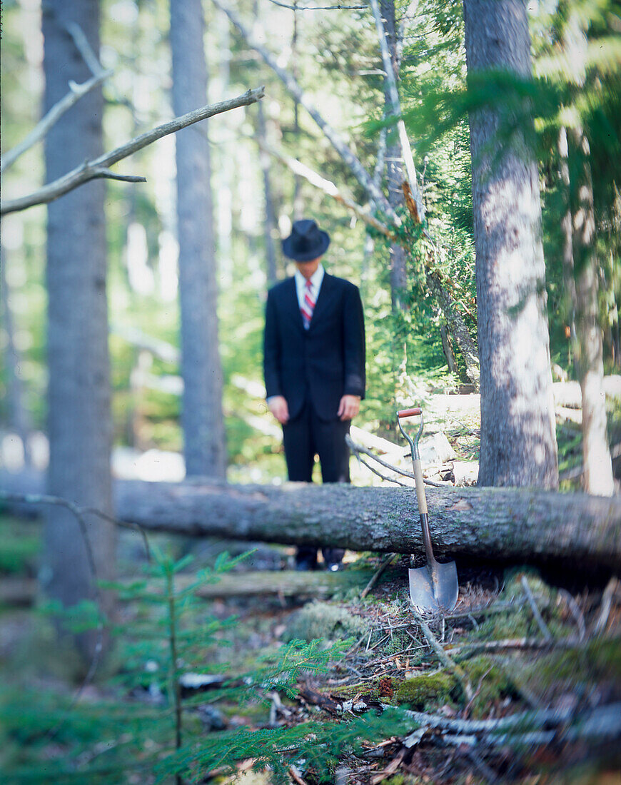 Man in suit and hat standing behind fallen tree and shovel in forest